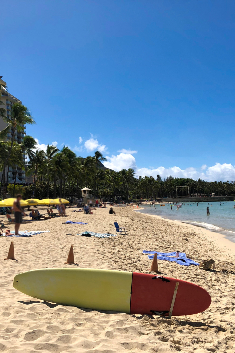 Surfboard resting on a sandy Oahu beach beside calm water and palm trees
