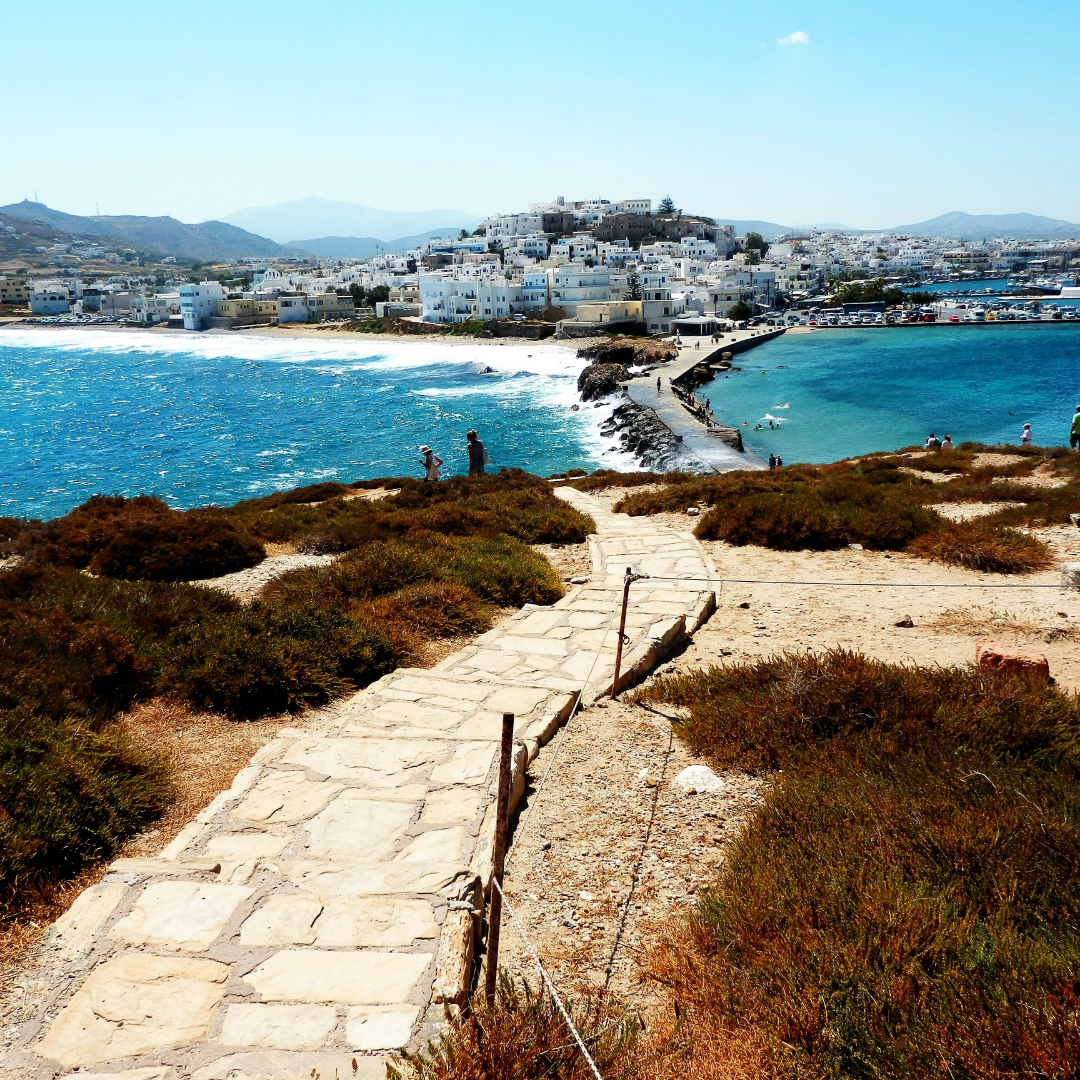 A stone pathway winding down a grassy hill leading toward a narrow causeway connecting to the white-washed buildings of Naxos town on a Greek island surrounded by blue ocean water.
