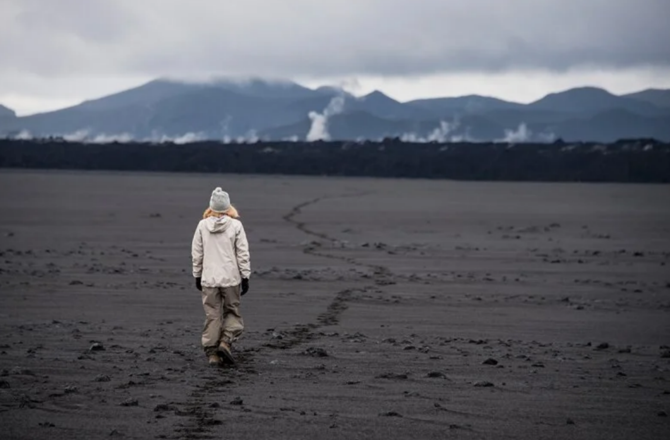 Hiker in a winter jacket walking across the expansive black lava sand plains of Holuhraun, Iceland under a cloudy sky.