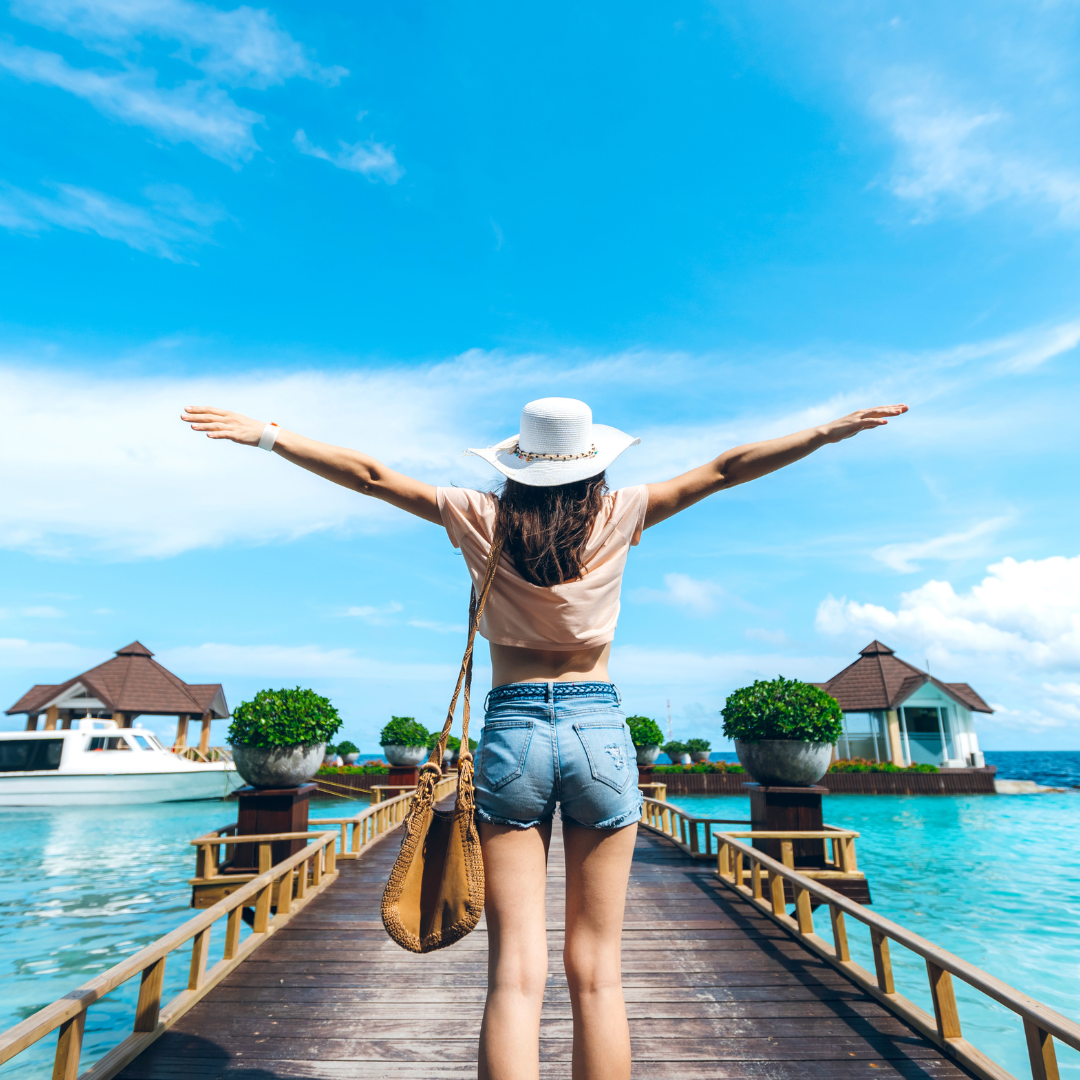 A joyful traveler celebrates her arrival at a luxury tropical island resort, standing on a pier with arms wide open under a blue sky.