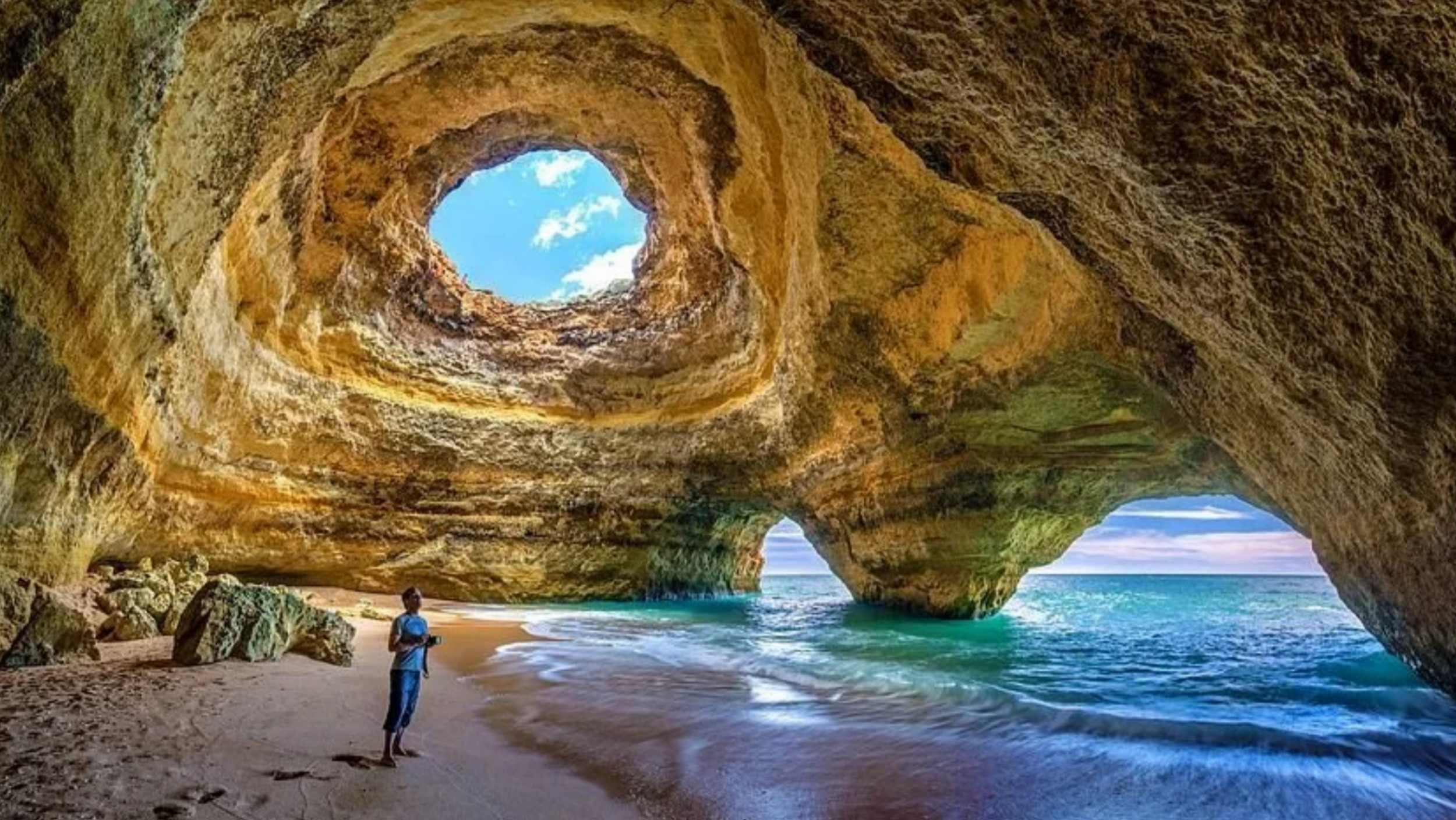 A person standing on the sandy beach inside the massive Benagil sea cave beneath a round natural skylight hole in the cavern ceiling in Portugal.
