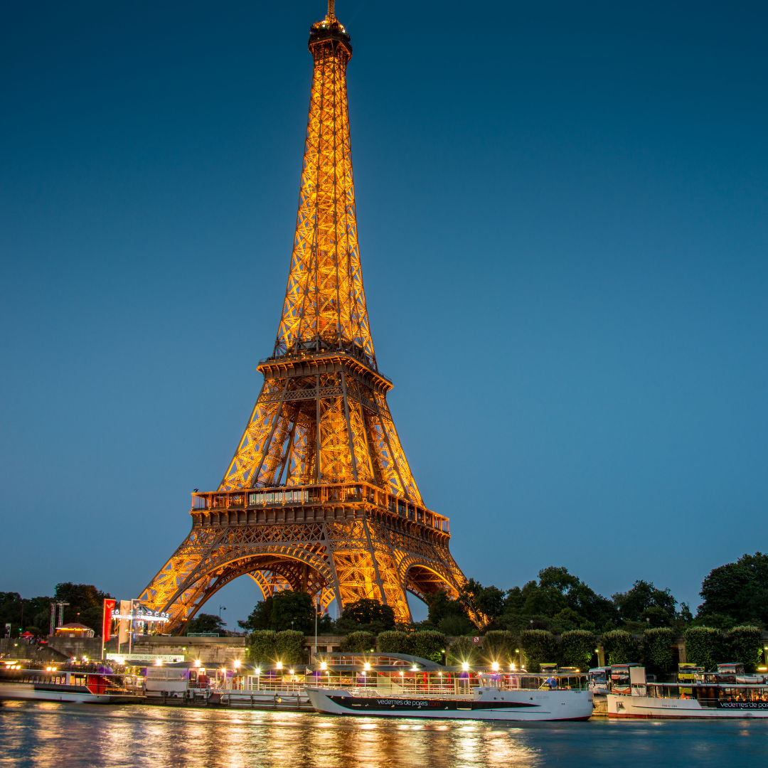 Glowing golden Eiffel Tower lit up at dusk with sightseeing boats cruising along the Seine River in Paris.