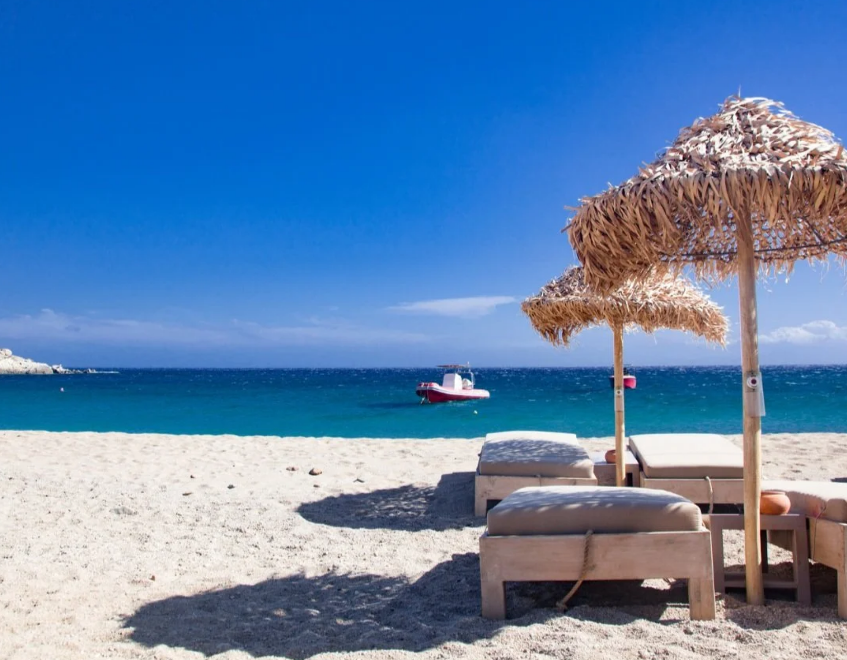 Mykonos beach in Greece with sun loungers and straw umbrellas overlooking clear turquoise waters and a boat in the Aegean Sea.