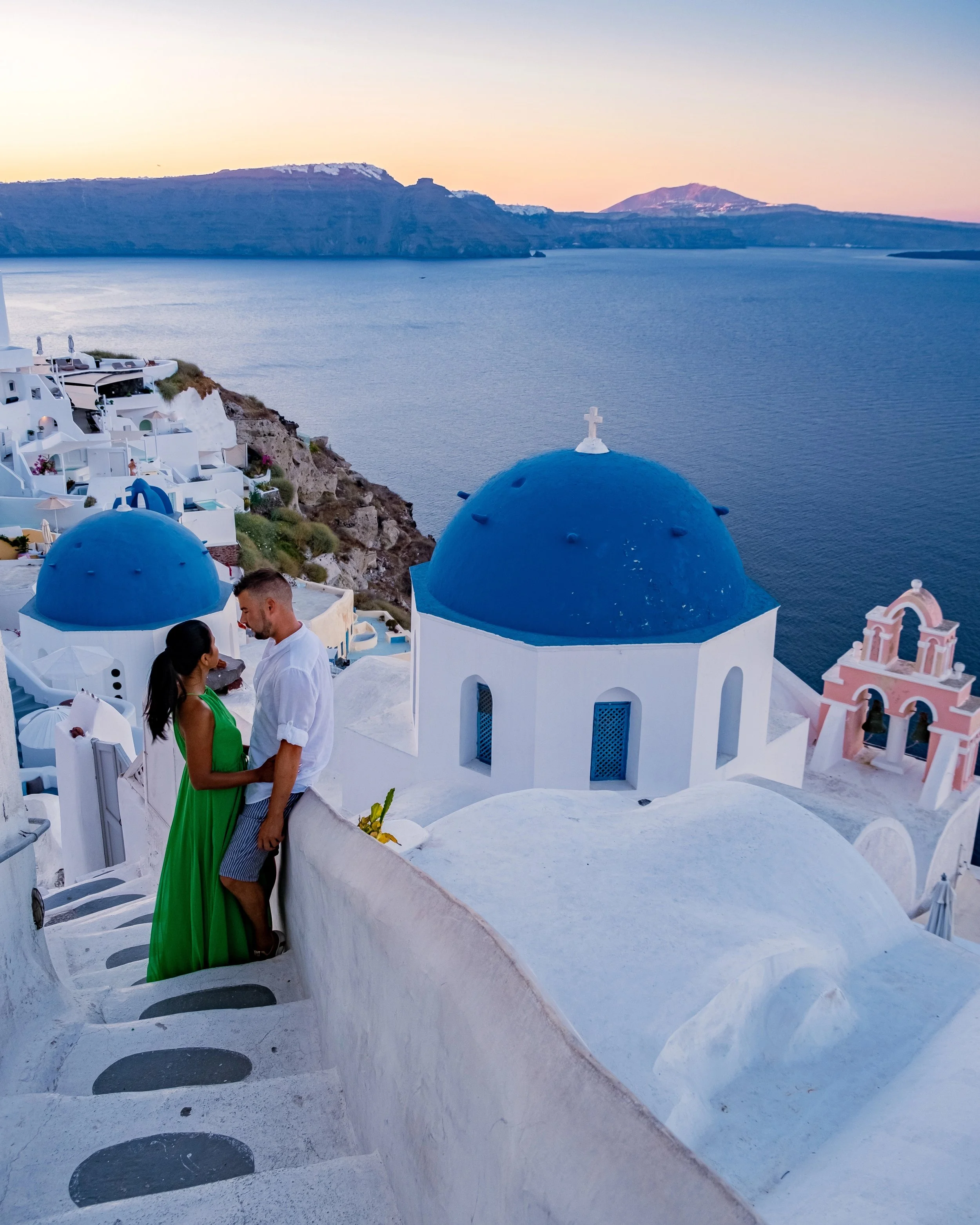 A romantic couple standing on a balcony overlooking the iconic blue-domed churches of Santorini, Greece, at sunset for a Honeymoon or Anniversary trip