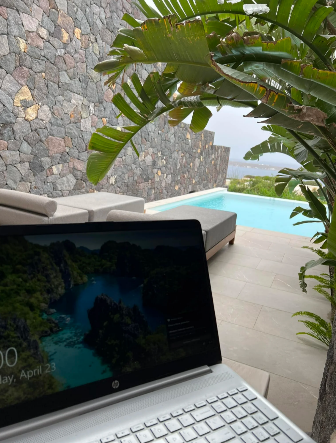 Close-up of an open laptop screen sitting on an outdoor patio next to a private plunge pool, stone wall, and tropical palm plant.