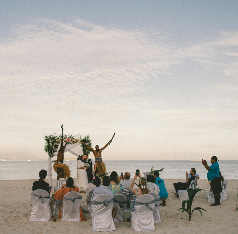 A bride and groom kissing under a floral arch during a tropical beach wedding ceremony with traditional island performers.