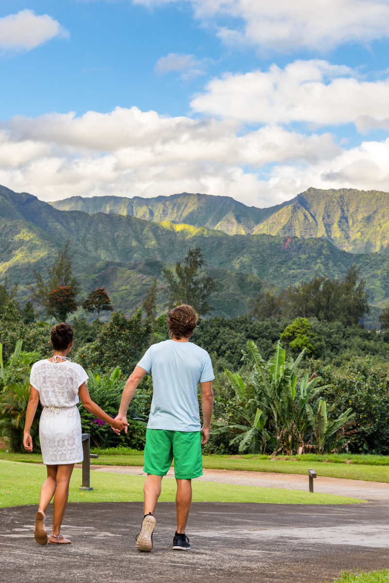 A couple walking hand-in-hand along an outdoor path facing dramatic, lush green volcanic mountains under a blue sky.