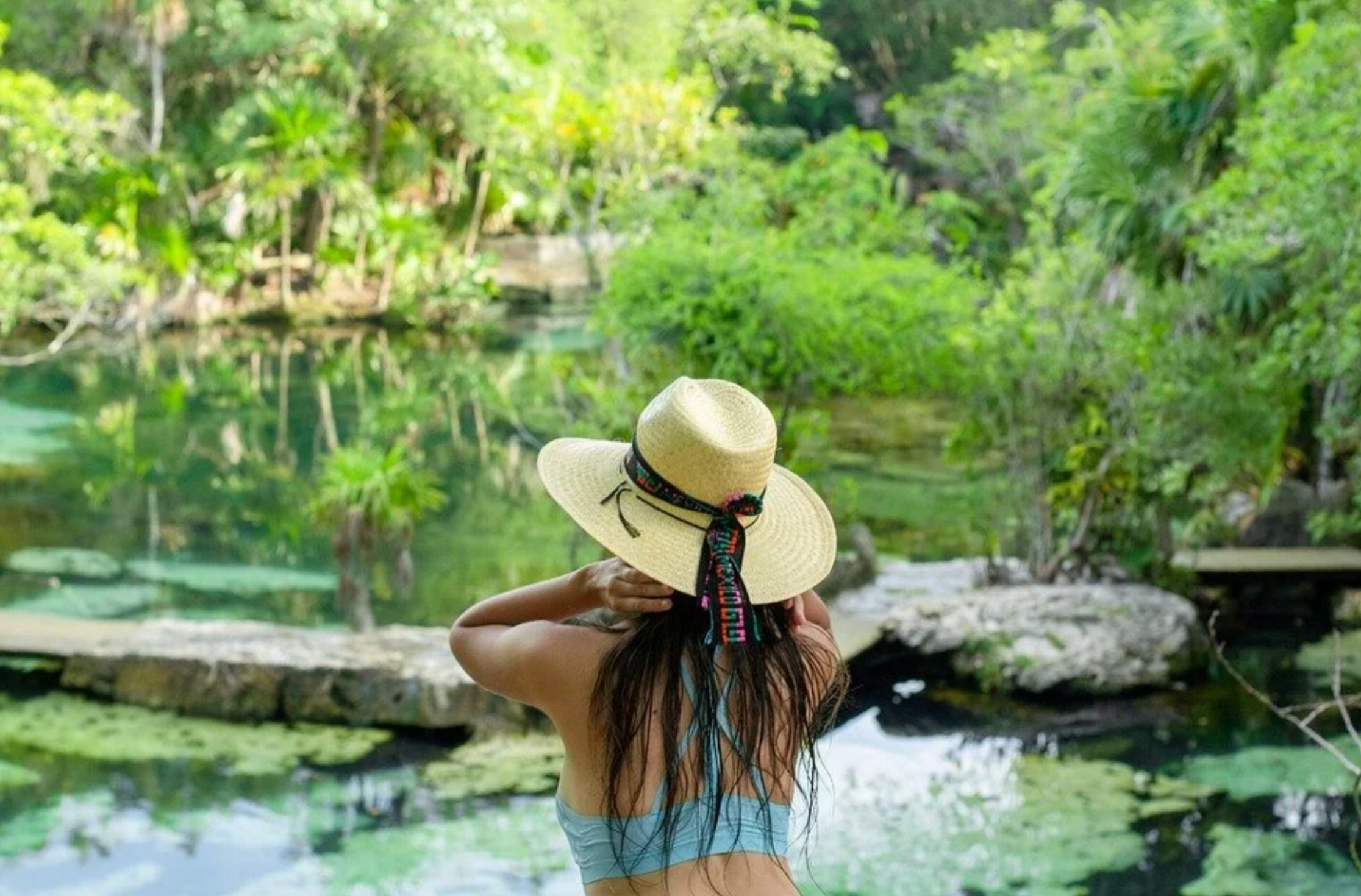 A woman in a straw hat from behind looking out over a calm, freshwater natural cenote surrounded by green jungle trees in Mexico.