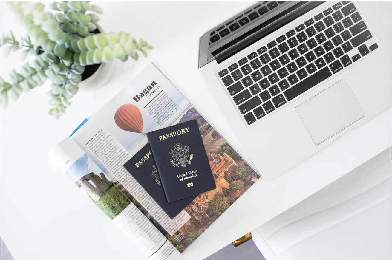 Flat lay of travel planning essentials including US passports, a travel magazine, and a laptop on a clean white desk.