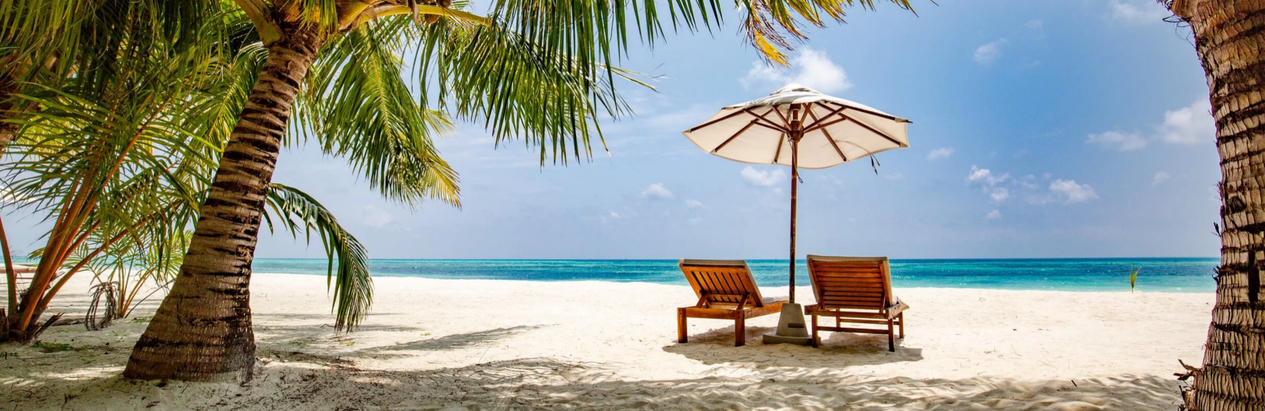Two wooden lounge chairs and a white umbrella under palm trees on a pristine white sand tropical beach facing blue ocean water.