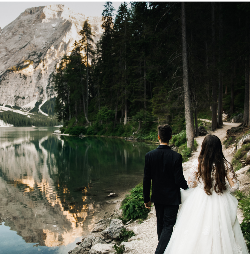 A bride and groom walking along a dirt path next to a crystal clear alpine lake with towering mountains in the background.