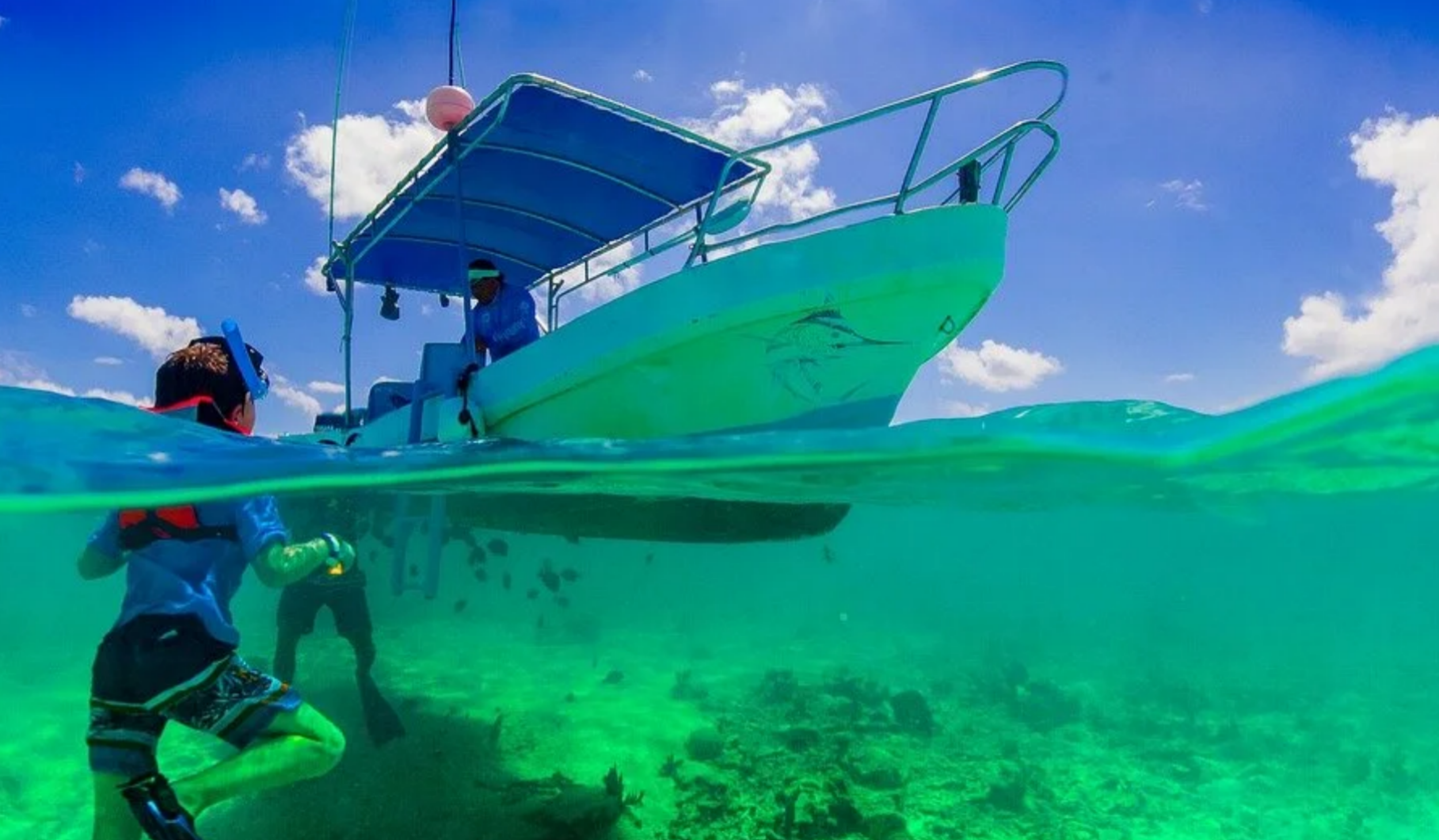 An over-under water shot showing a young snorkeler in a life vest in clear green ocean water beneath a small blue canopy boat.