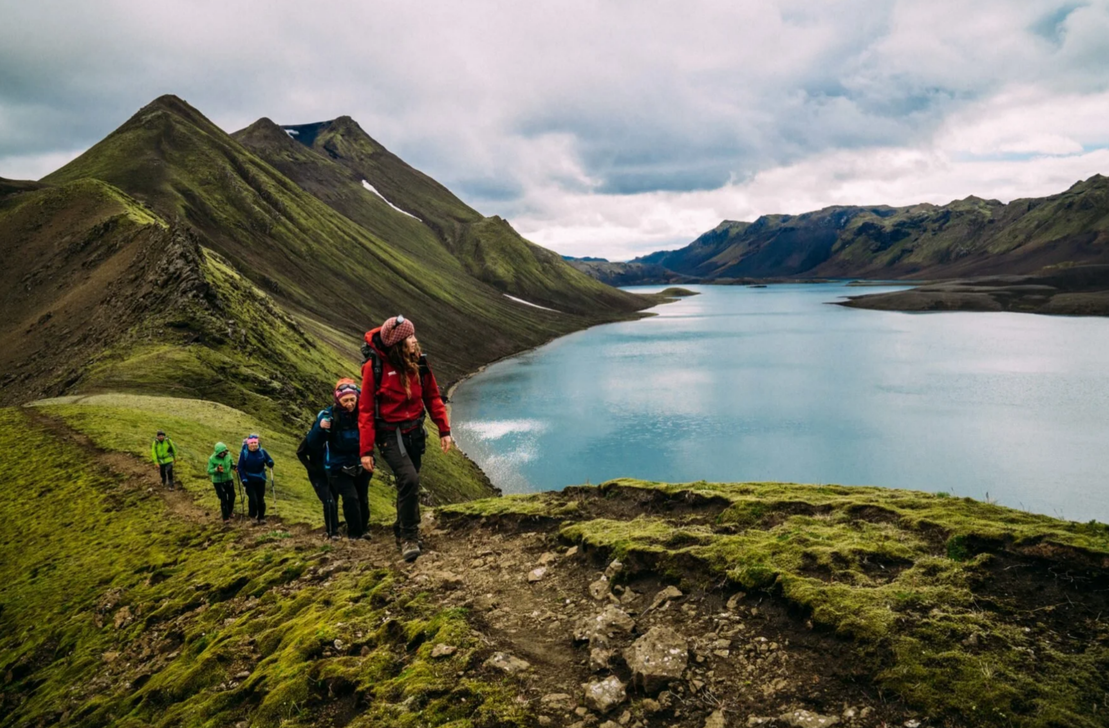 A line of hikers with backpacks trekking along a mossy green mountain ridge next to a large, calm blue volcanic lake in Iceland.