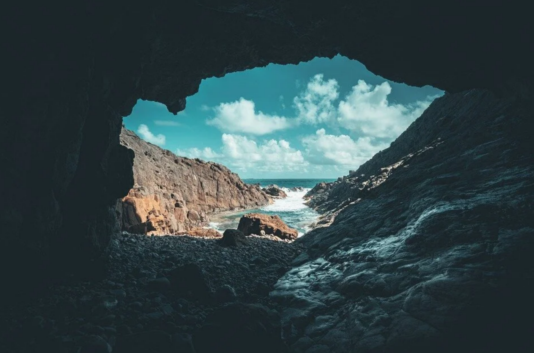 Looking out from the dark interior of a coastal sea cave toward crashing white ocean waves and rocky cliffs under a blue sky in Puerto Rico.