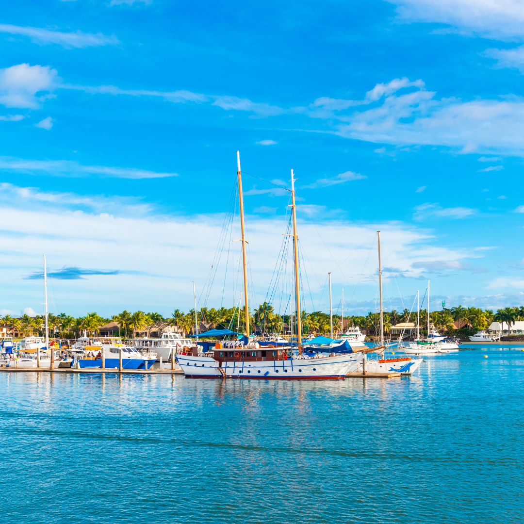 Luxury yachts and large white masted sailboats docked at a tropical marina harbor under a blue sky with light clouds.