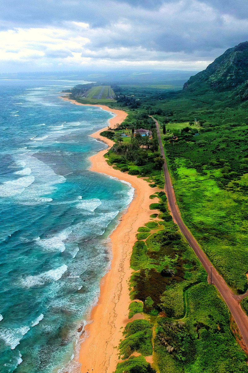 Aerial view of a long golden sand beach and turquoise breaking ocean waves running alongside a road and steep green mountains.