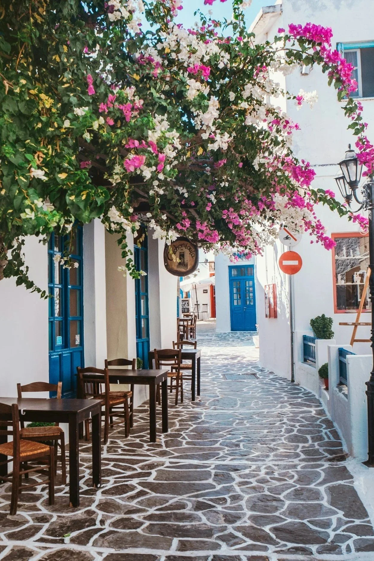 A charming cobblestone street in a Greek village with white-washed buildings, blue doors, and blooming pink bougainvillea flowers.