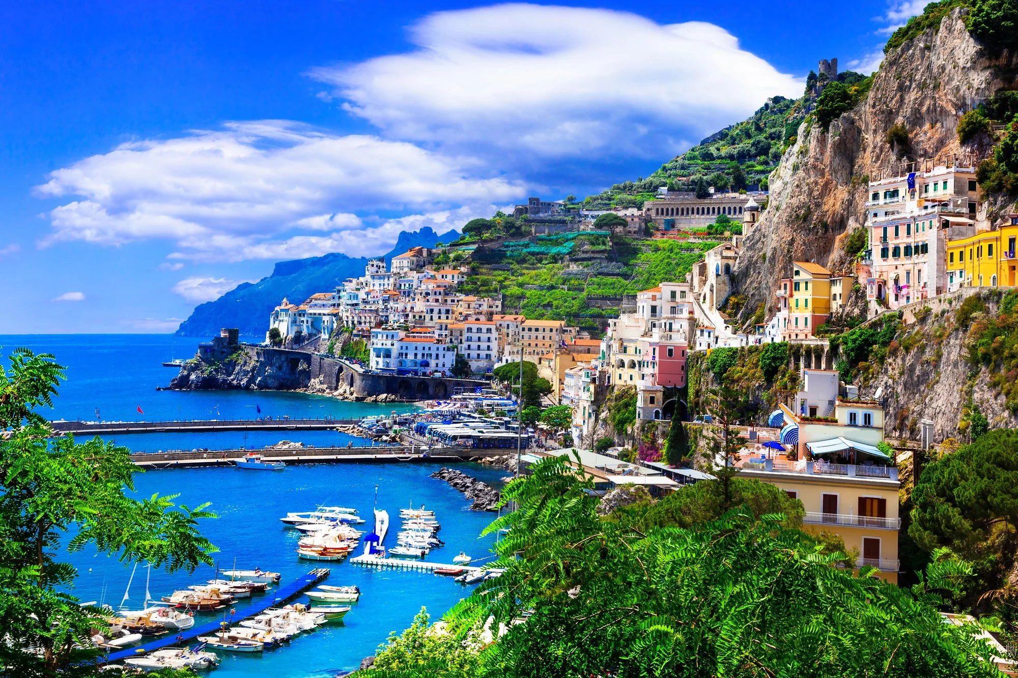 Aerial view of the Amalfi Coast in Italy showing colorful cliffside buildings, a blue harbor with boats, and lush green mountains overlooking the Mediterranean Sea.