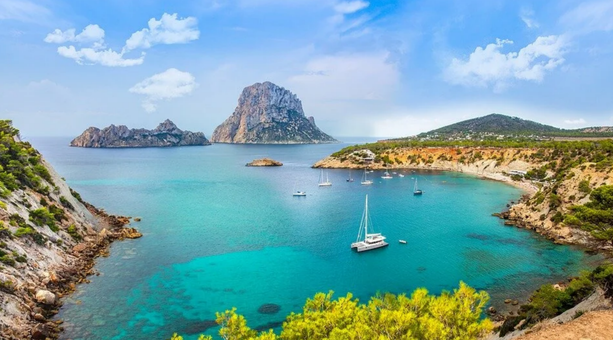 High-angle view of a turquoise cove with sailboats anchored in the bay looking out at the majestic Es Vedrà rock island in Ibiza, Spain.