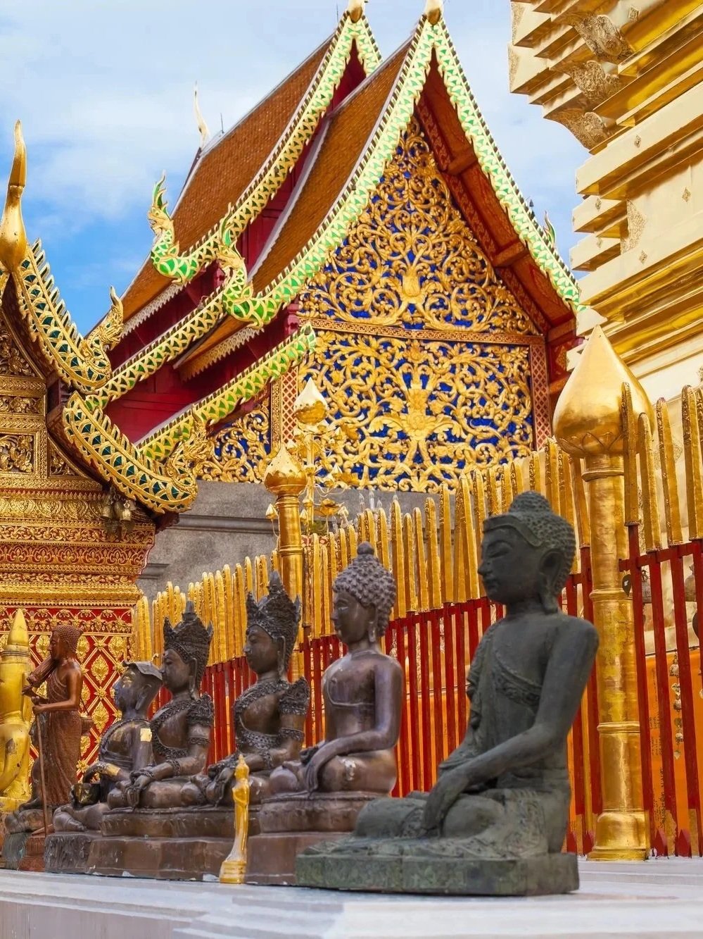 A row of ancient bronze Buddha statues in front of a stunning gold and red ornate Buddhist temple in Thailand, illustrating cultural immersion tours by Lavishly Travel.