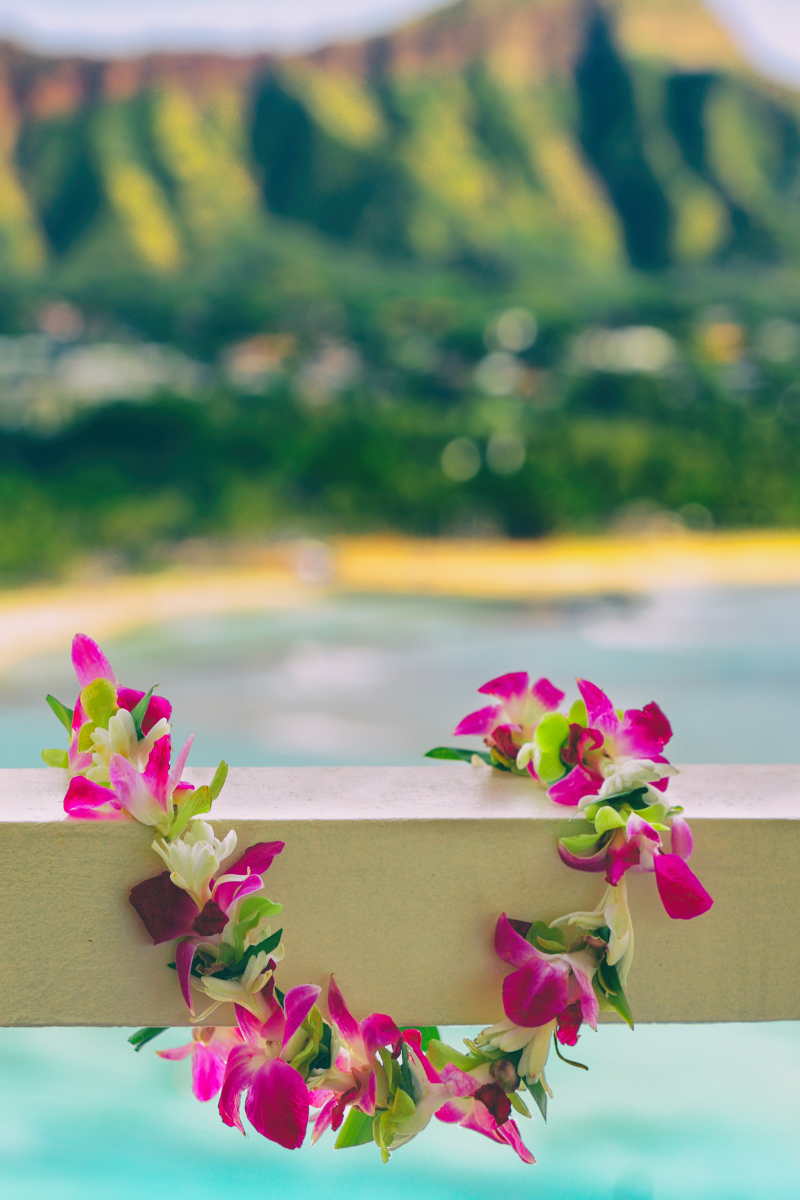 A fresh purple and white orchid lei draped over a balcony railing with blurred tropical mountains in the background.