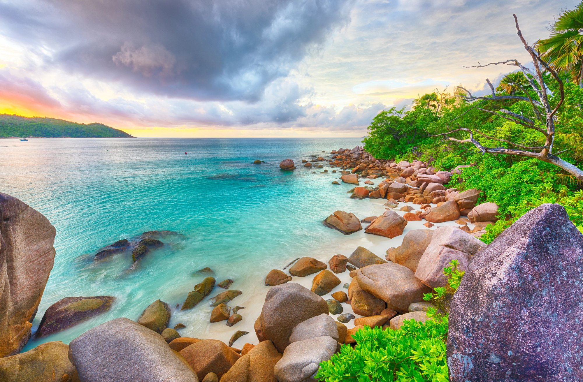 Turquoise ocean water meeting a white sand beach with large orange-tinted granite boulders and lush greenery under a dramatic sky in Australia.