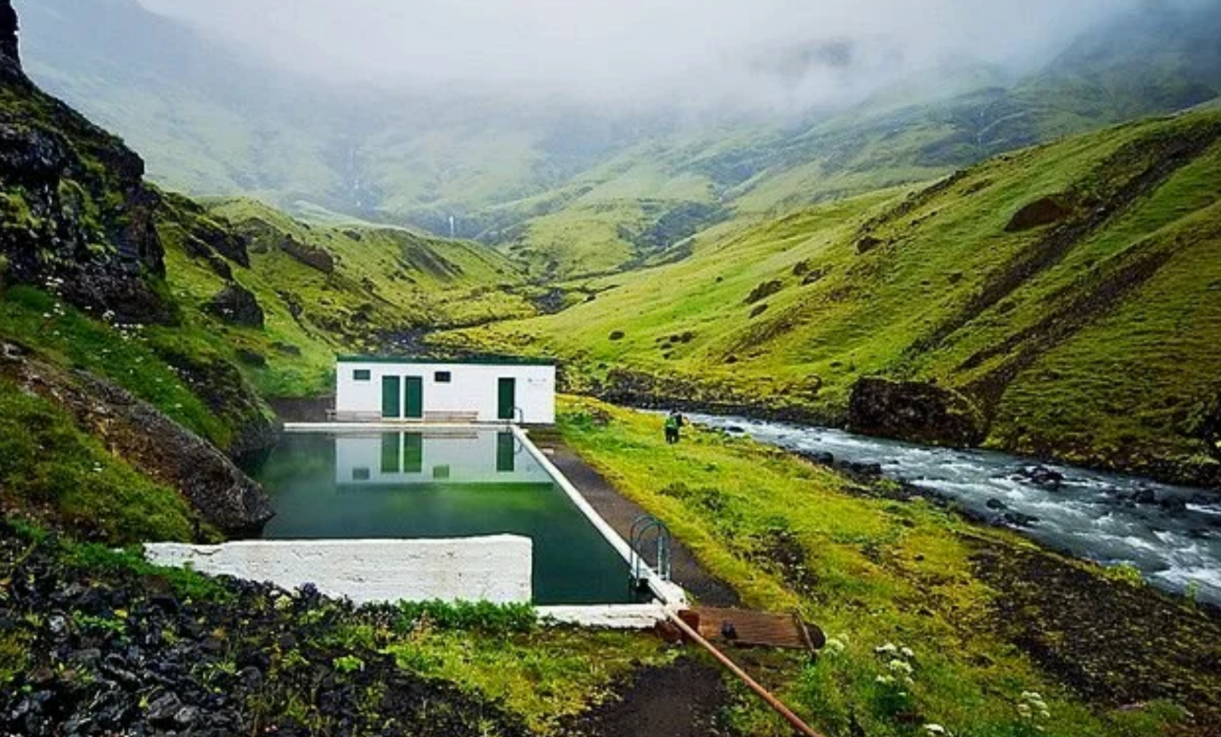 The historic Seljavallalaug geothermal pool and changing room situated in a deep, vibrant green mountain valley next to a rushing river.