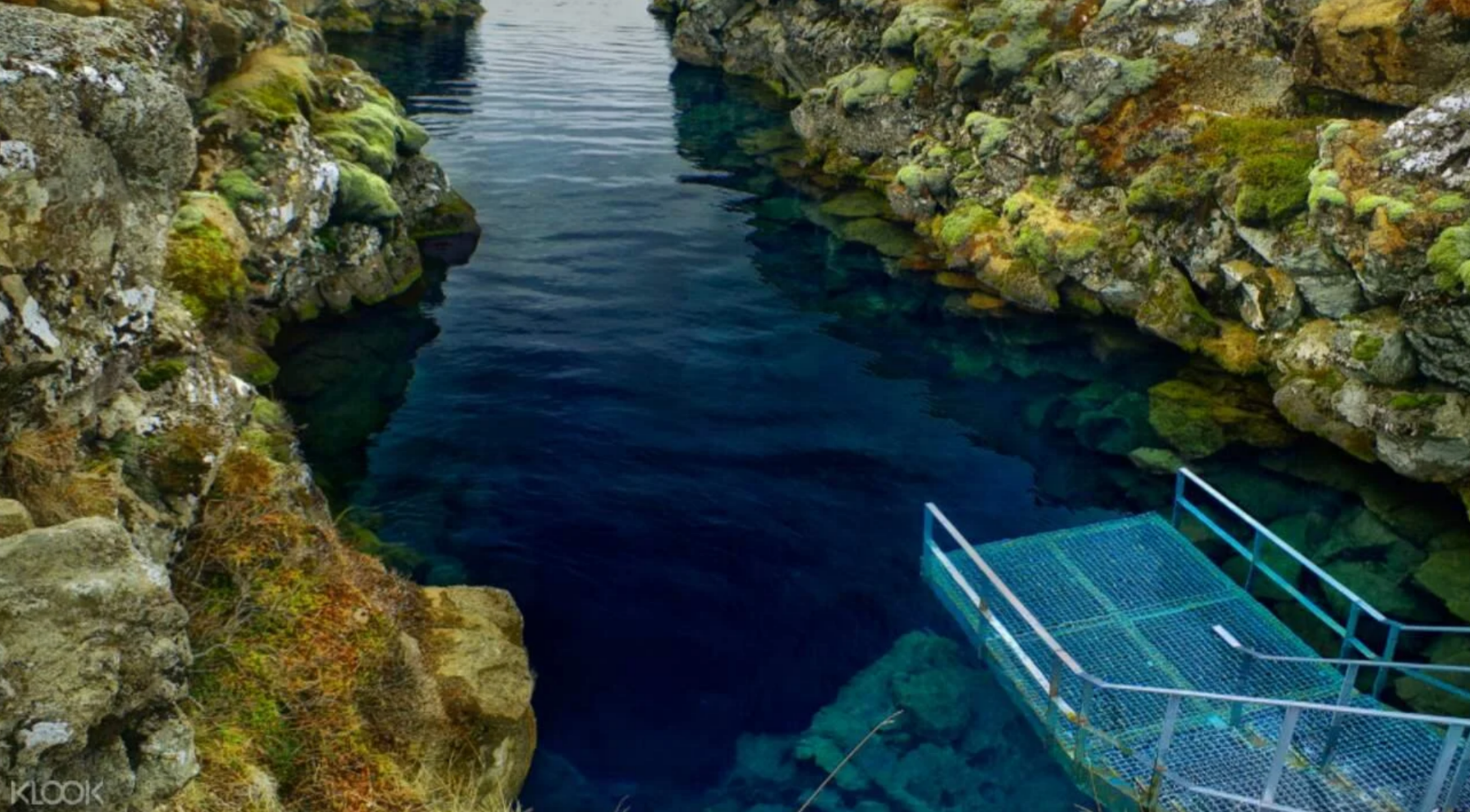 A metal diving platform leading into the vibrant, crystal-clear deep blue water of the Silfra Fissure between mossy rock walls.