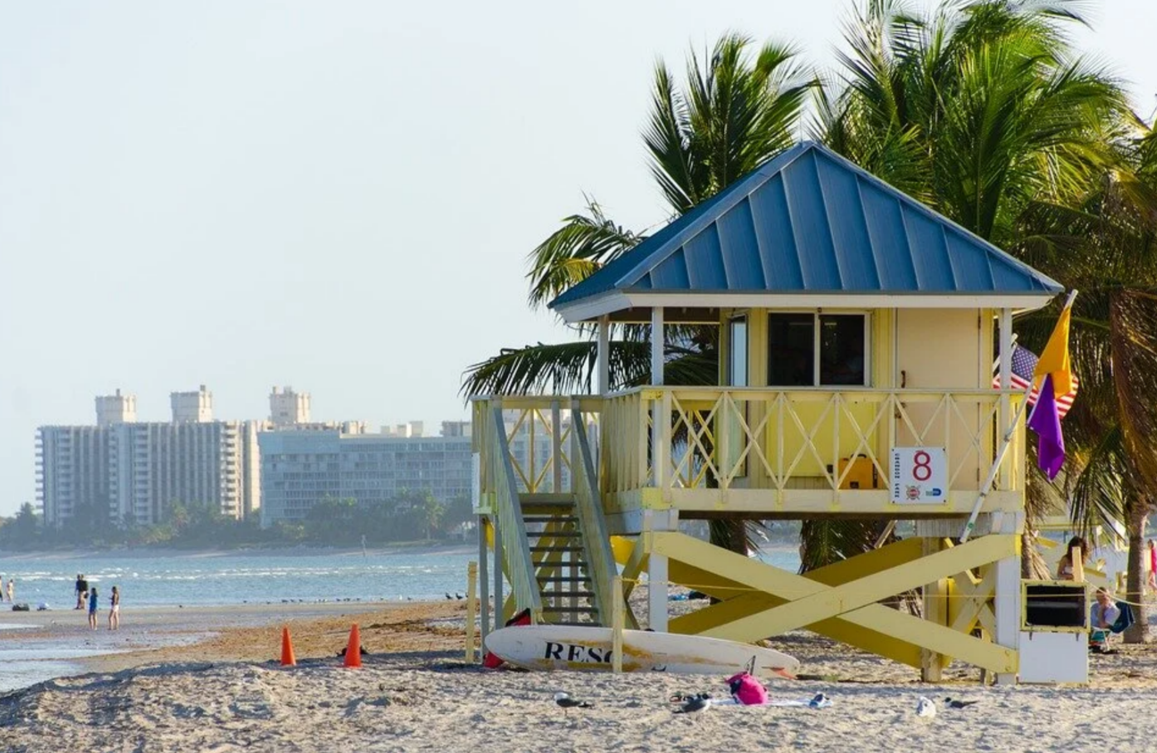 An iconic yellow and blue wooden lifeguard station on the sandy beach in Miami, Florida with palm trees and high-rise hotel buildings in the background.