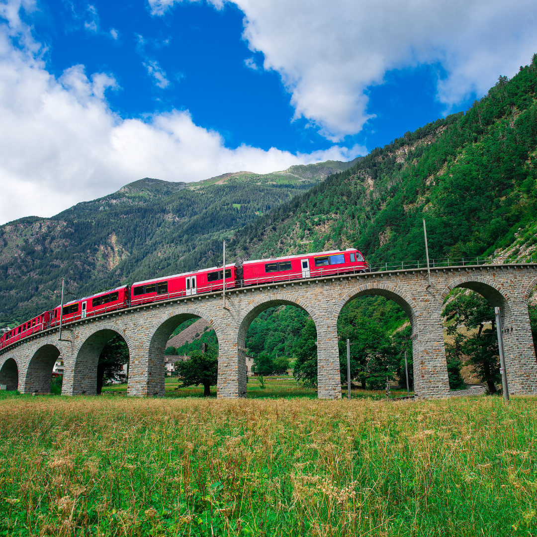 The bright red Bernina Express passenger train crossing a historic stone-arched viaduct framed by green hills and Swiss Alpine mountains.