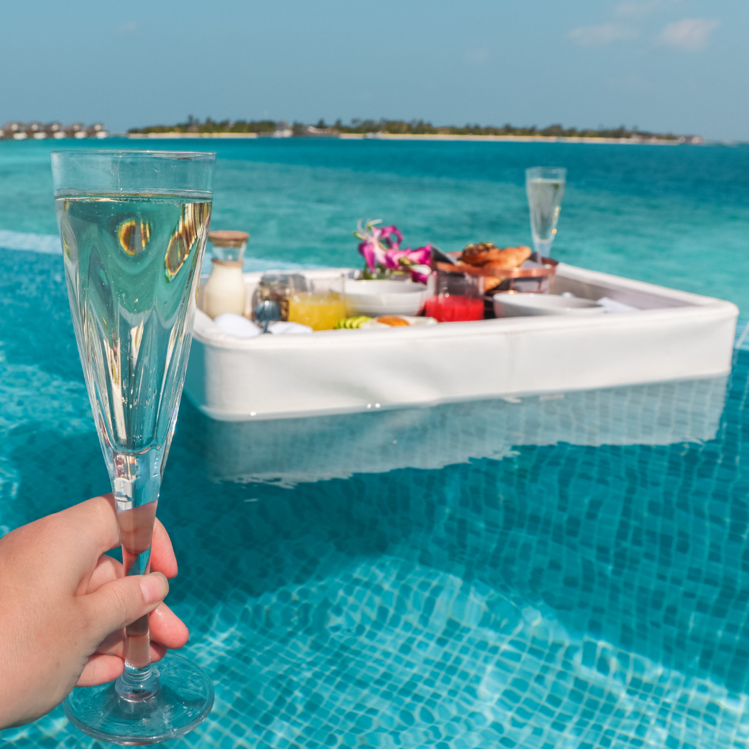 hand holding a glass of champagne in a crystal-clear blue infinity pool with a luxury floating breakfast tray filled with juices and pastries in the Maldives.