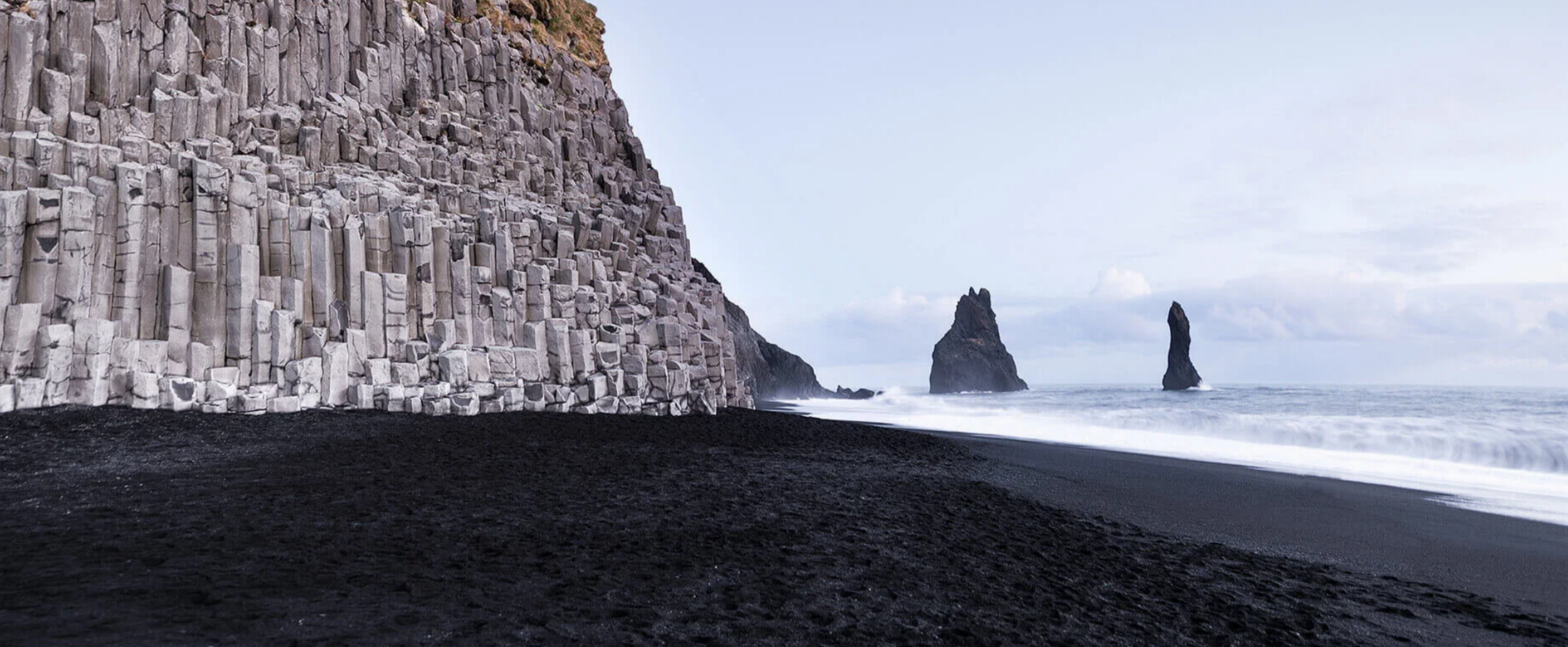 The geometric basalt column cliffs on Reynisfjara black sand beach with the Reynisdrangar sea stacks visible in the crashing ocean waves.