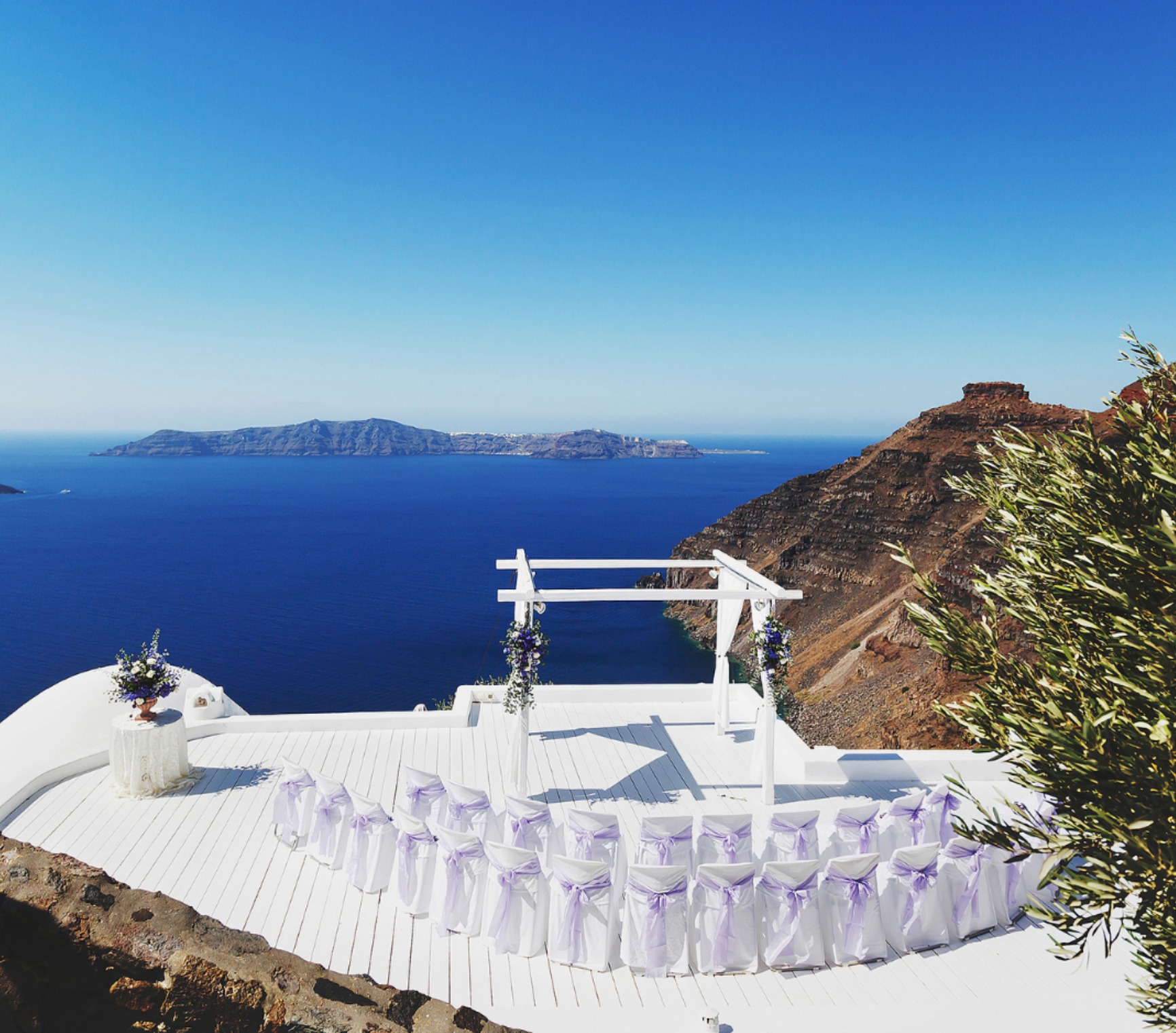 A white wedding ceremony terrace overlooking the deep blue Aegean Sea and volcanic islands in Santorini, Greece.