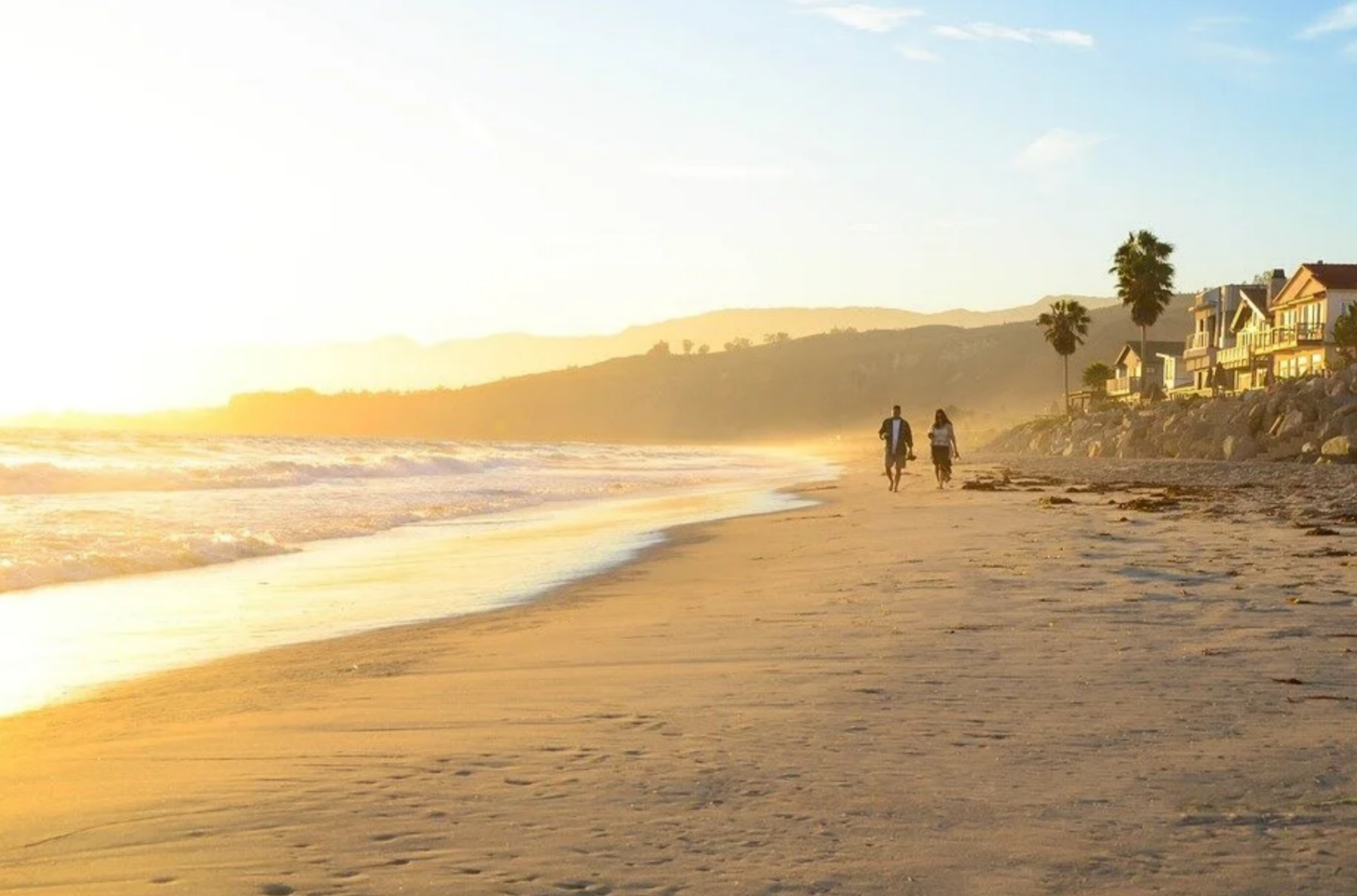 A couple walking together along the shoreline of a wide sandy beach bathed in warm golden sunset light near Malibu and Los Angeles coastal homes.