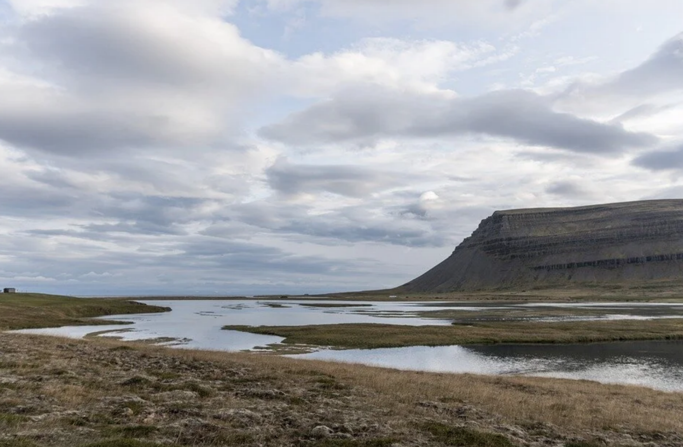 A calm, flat water estuary river winding through grassy wetlands toward a flat-topped mountain cliff in the Westfjords, Iceland.