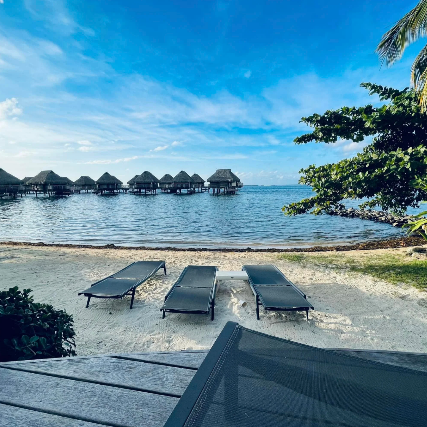 Lounge chairs sitting on a white sandy beach in front of clear turquoise water and a line of overwater bungalows.