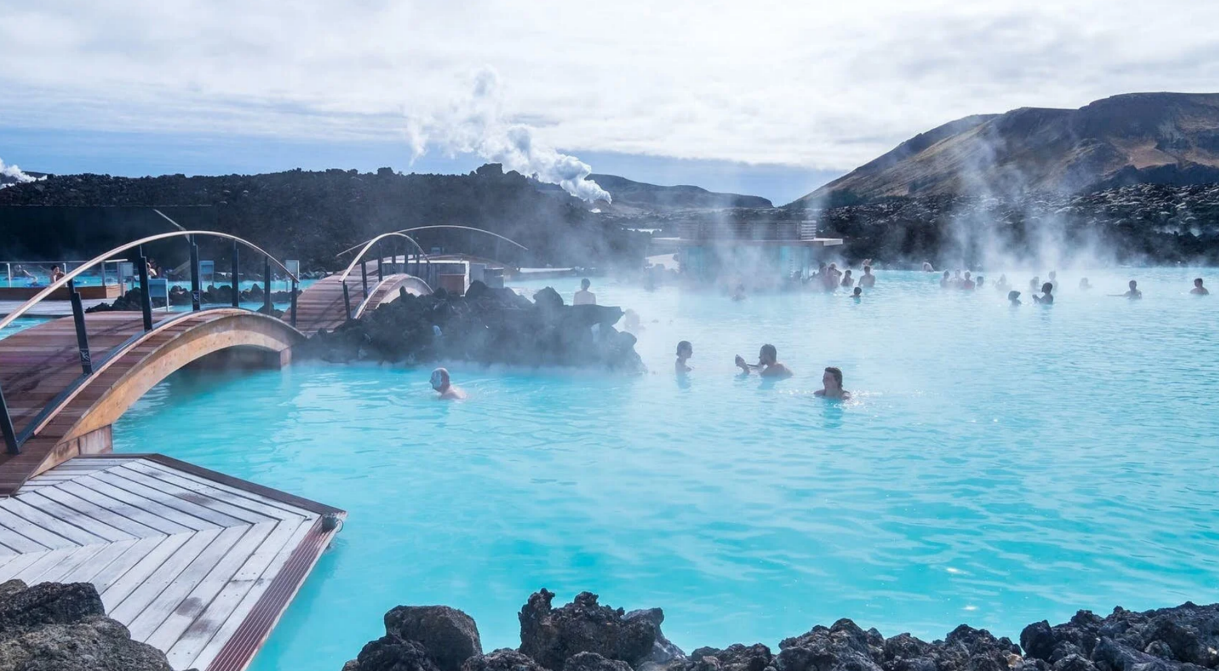 People swimming and relaxing in the steaming, bright turquoise geothermal water of the Blue Lagoon in Iceland near a wooden footbridge.