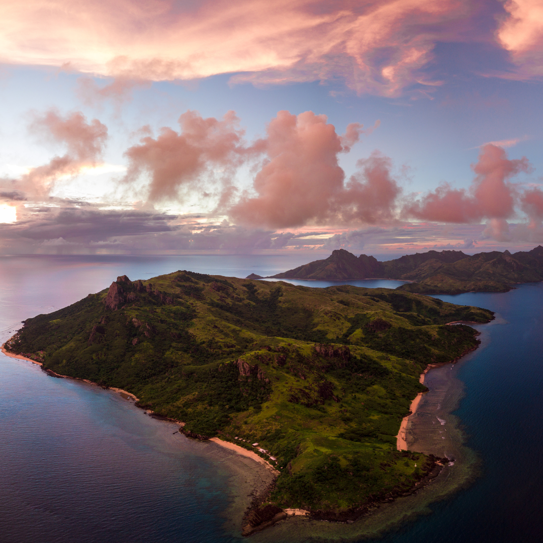Spectacular aerial view of a green volcanic tropical island in the ocean under a dramatic pastel pink and orange sunset sky.