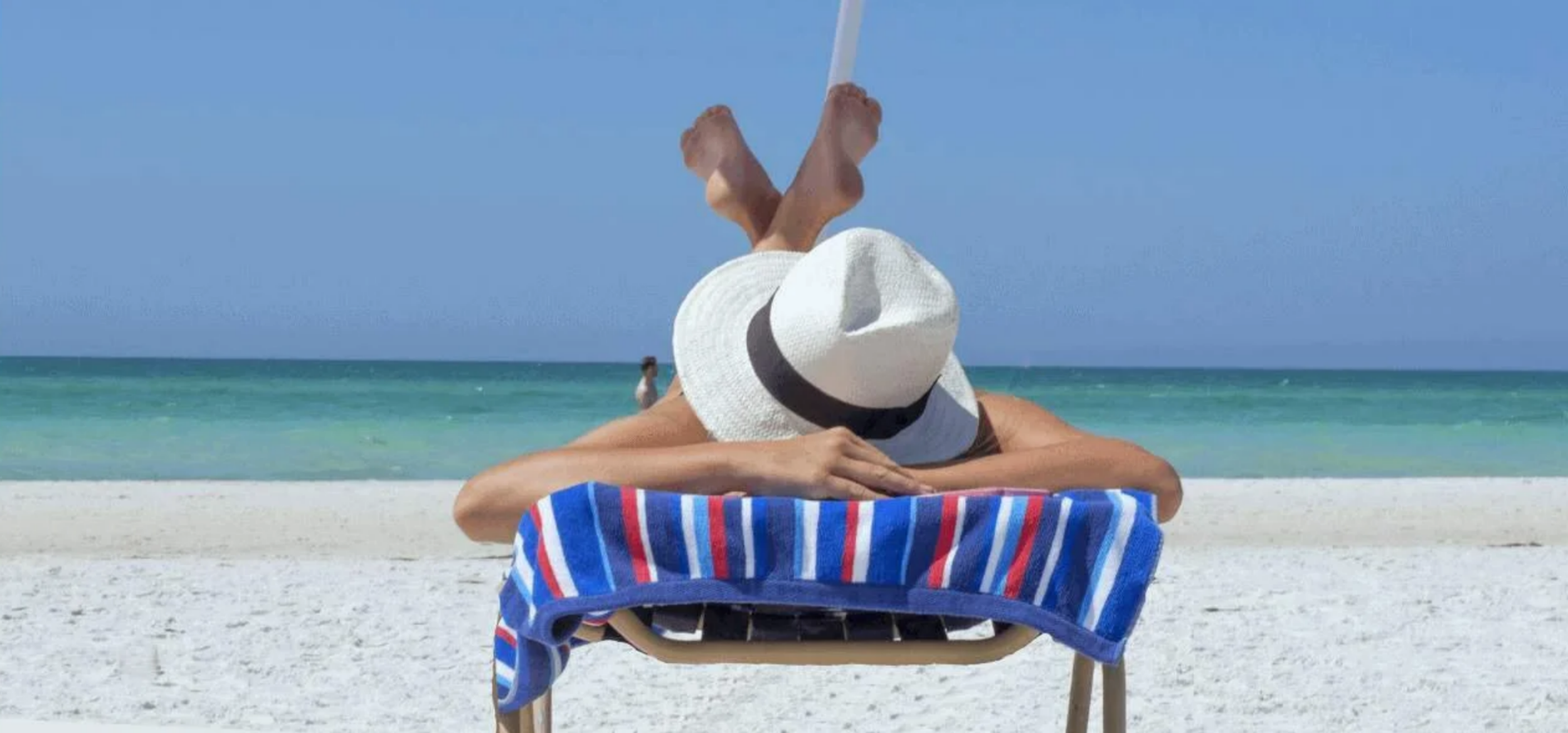 person lounging back-first on a beach chair under an umbrella wearing a white sun hat facing the turquoise sea.