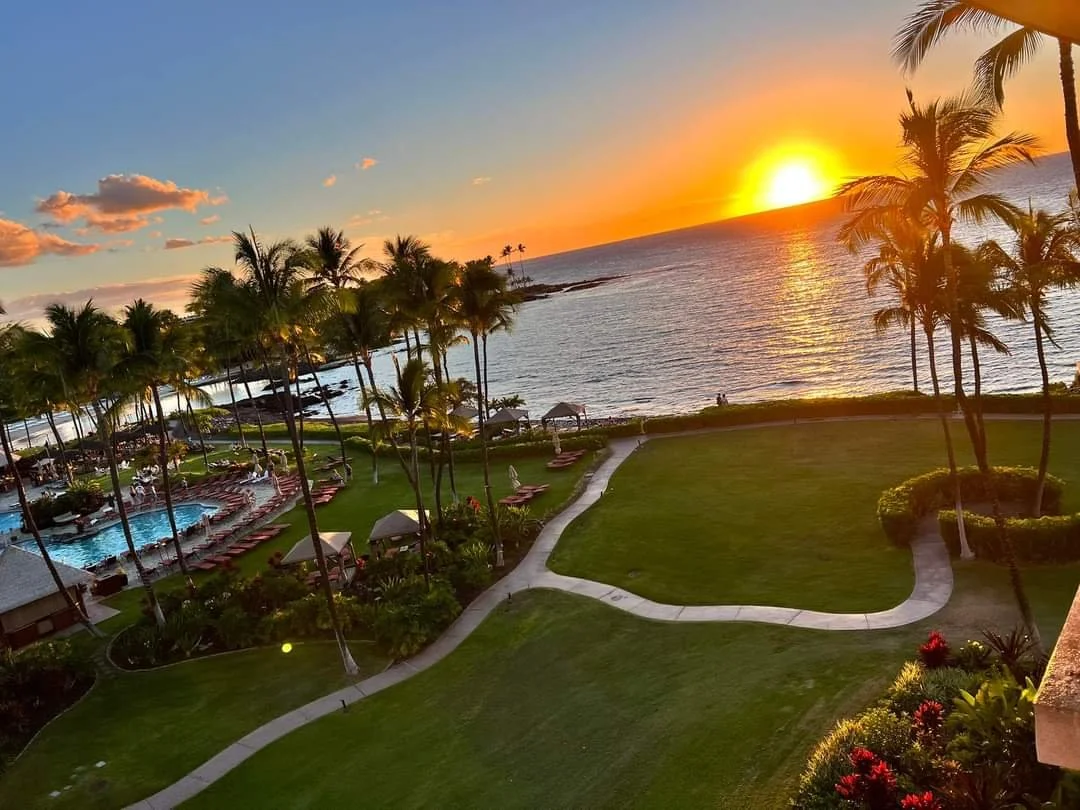 Sunset over the ocean from a resort lanai overlooking palm trees, manicured lawns, and the swimming pool at the Fairmont Orchid on Hawaii's Kohala Coast.