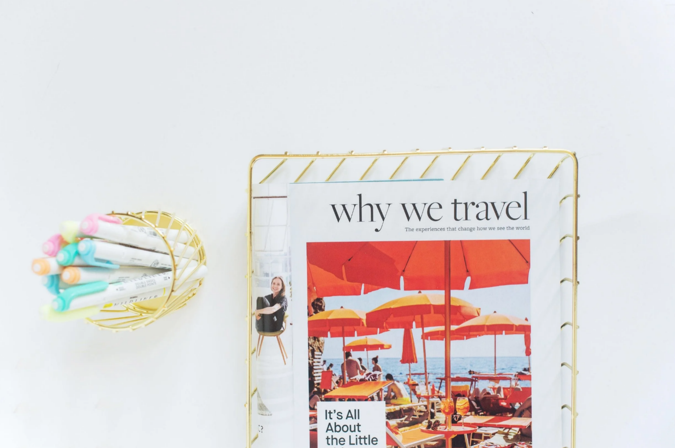 A flat lay of a magazine showing a beach scene with orange umbrellas next to a gold wire holder filled with pastel markers.