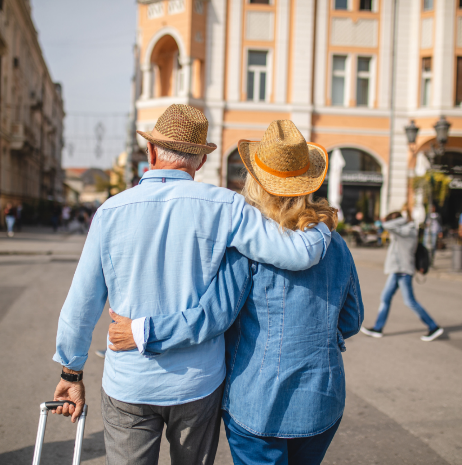 Rear view of a senior couple in straw hats walking arm-in-arm down a European city street pulling rolling luggage.