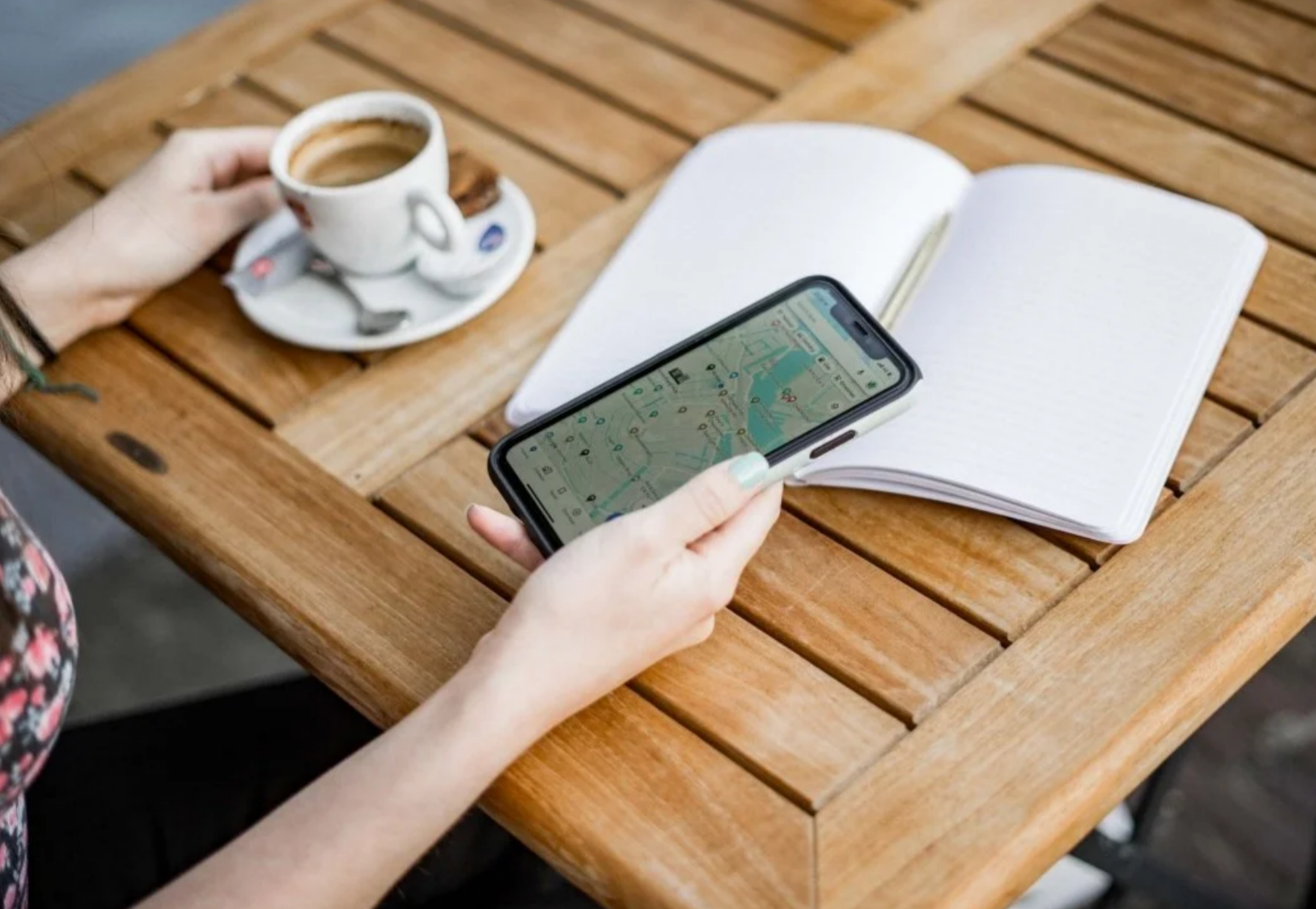 Person holding a smartphone displaying a digital map while sitting at a wooden cafe table next to an espresso cup and an open blank journal.