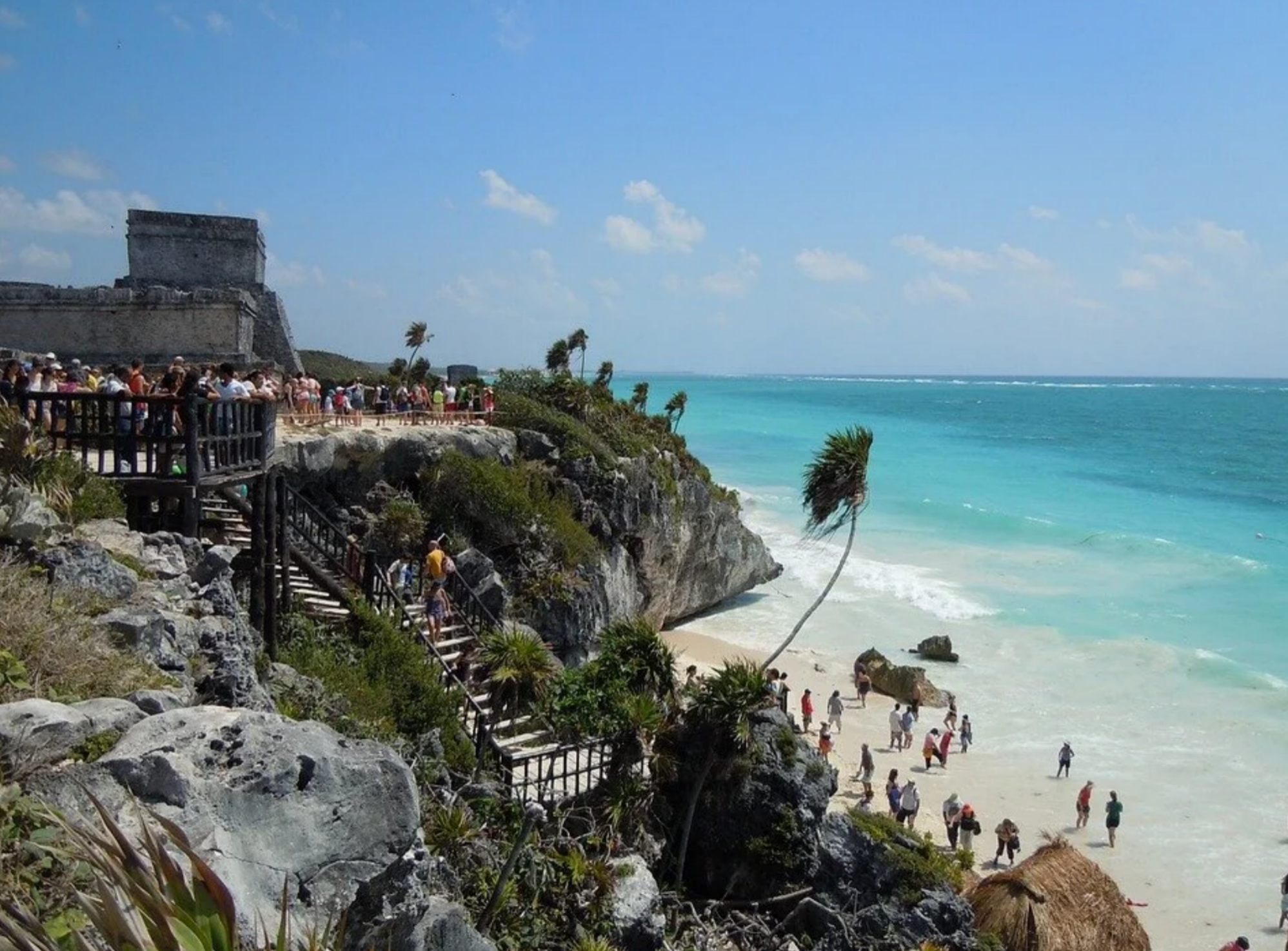 Historical stone Mayan ruins atop high coastal cliffs with visitors overlooking a sandy beach and vivid turquoise sea in Tulum, Mexico.