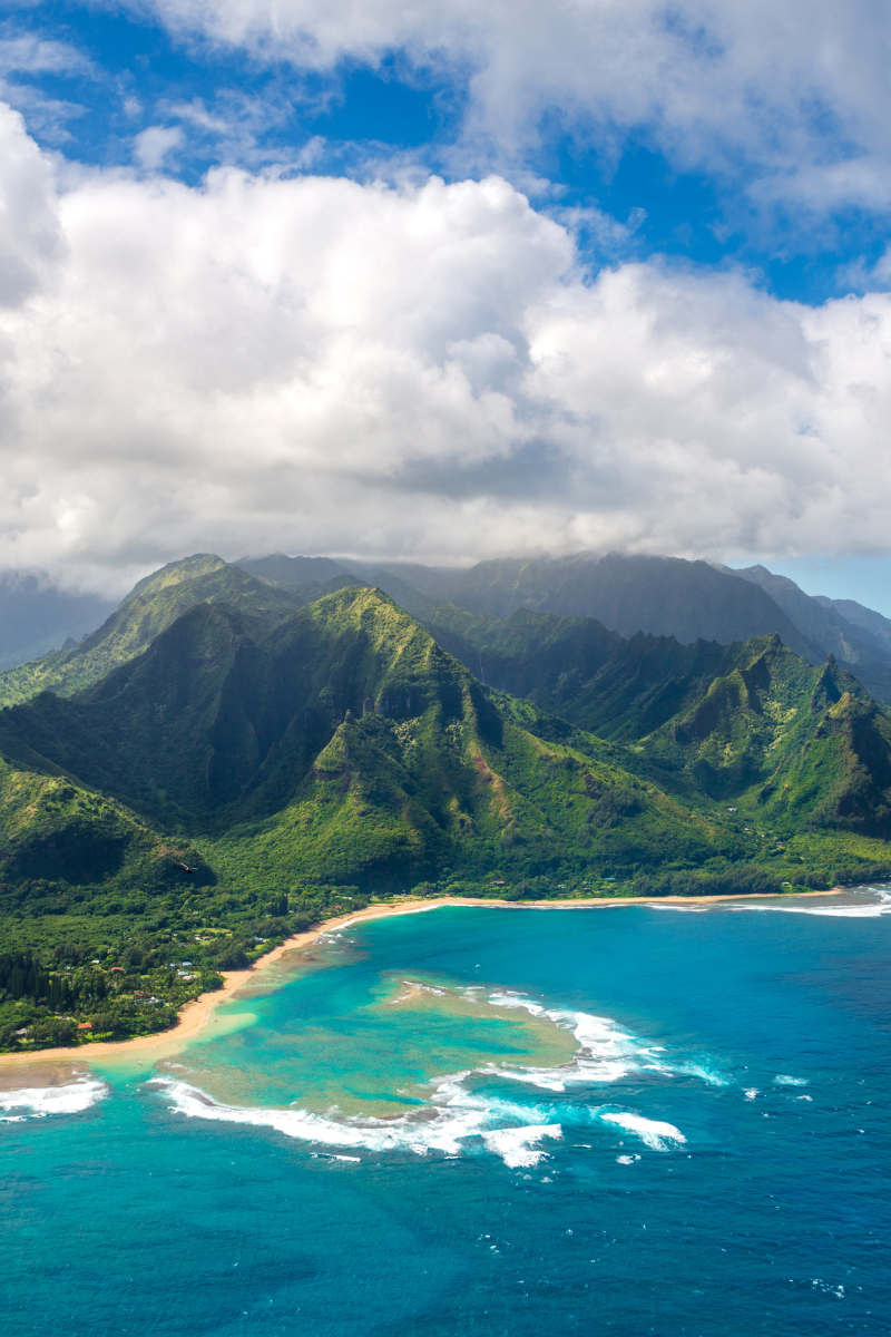 Aerial perspective of dramatic green mountain peaks and emerald valleys meeting a curved golden beach and turquoise ocean in Kauai.