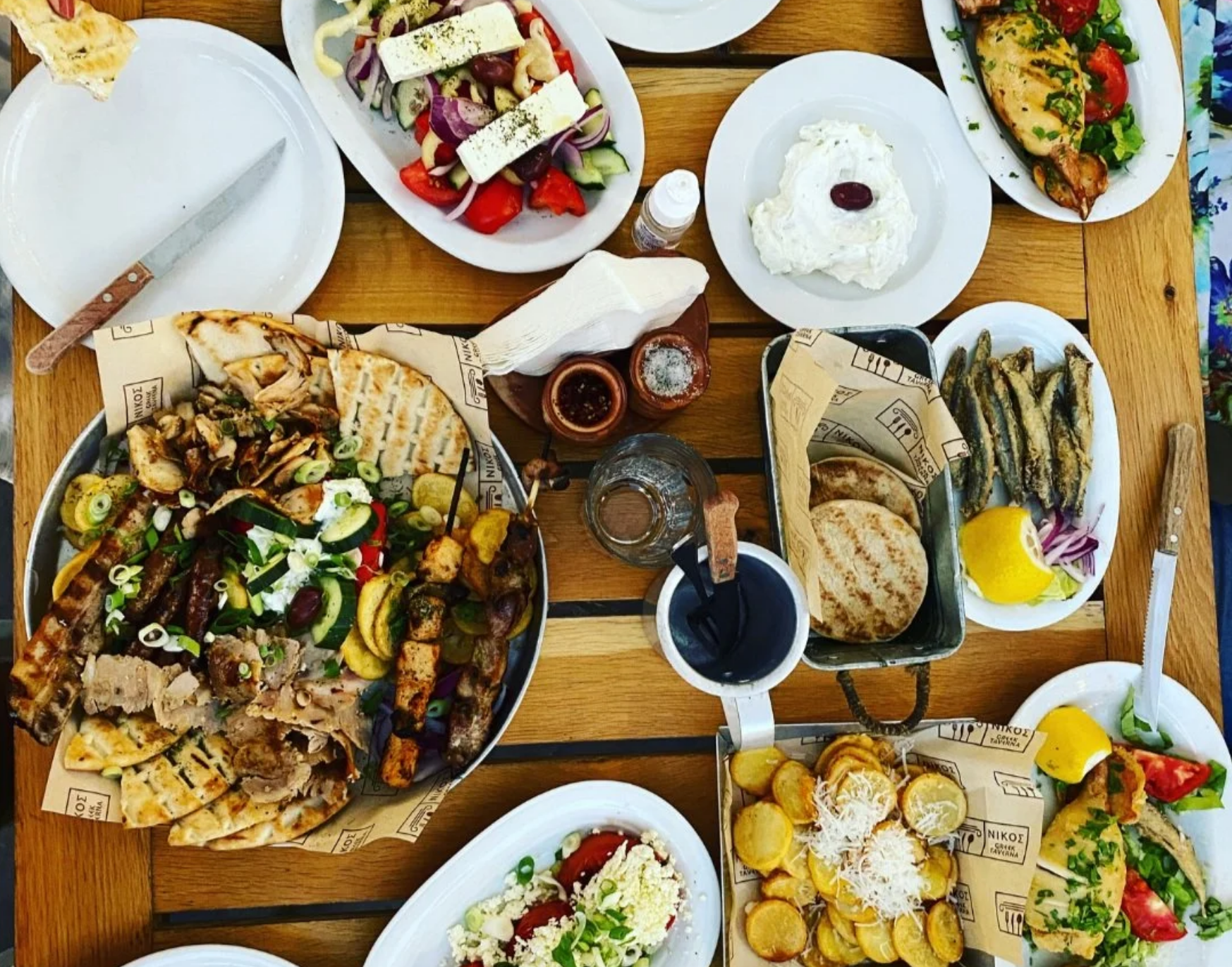 Overhead view of a shared meal with salad, bread, dips and grilled dishes arranged on a wooden table.