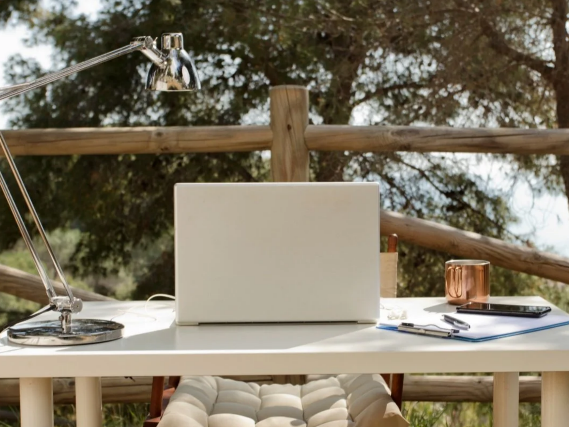 Minimalist white desk set up outdoors with a closed laptop, desk lamp, notebook, and copper mug against a wooden fence and tree background.