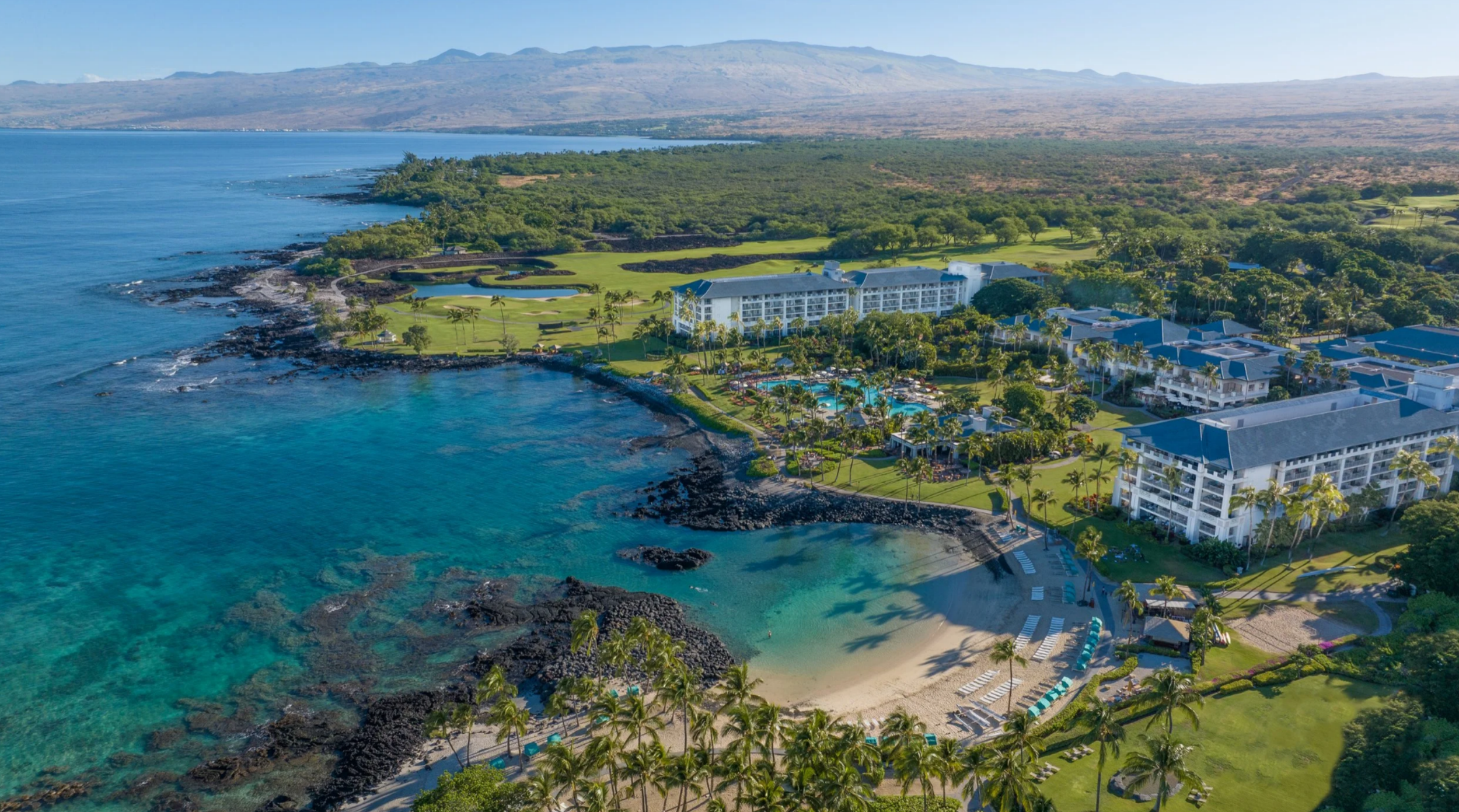 Panoramic aerial landscape showing a beachfront resort nestled along a clear blue ocean cove, dark lava rock shores, sandy beach area, and sprawling mountain terrain in the background.