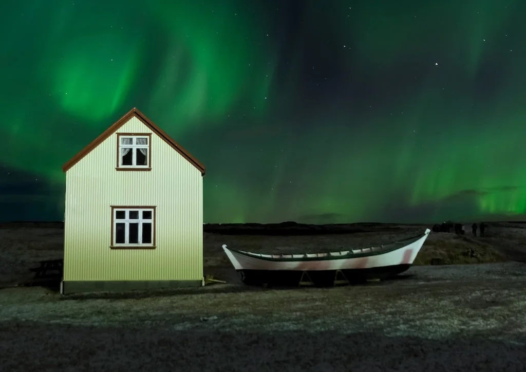 A small two-story corrugated iron house next to a traditional beached wooden boat under a massive, swirling green Aurora Borealis night sky.