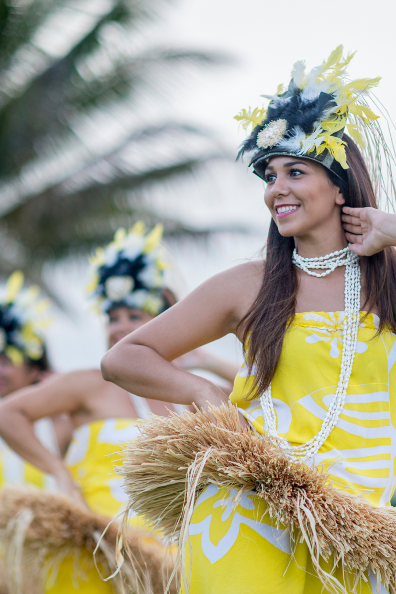 A Hawaiian dancer in a bright yellow floral outfit, shell necklace, and feathered headpiece performing outdoors.